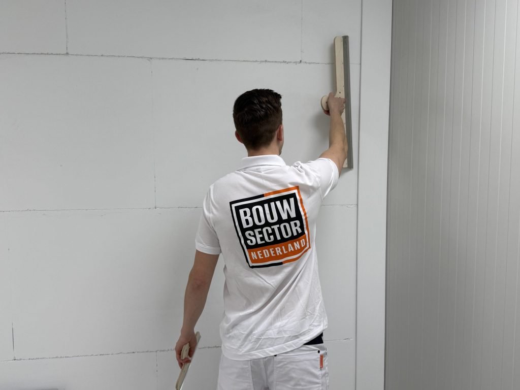 Stukadoor met het logo van Bouwsector Nederland op zijn rug trekt hoog op de wand een lange spaan omhoog
