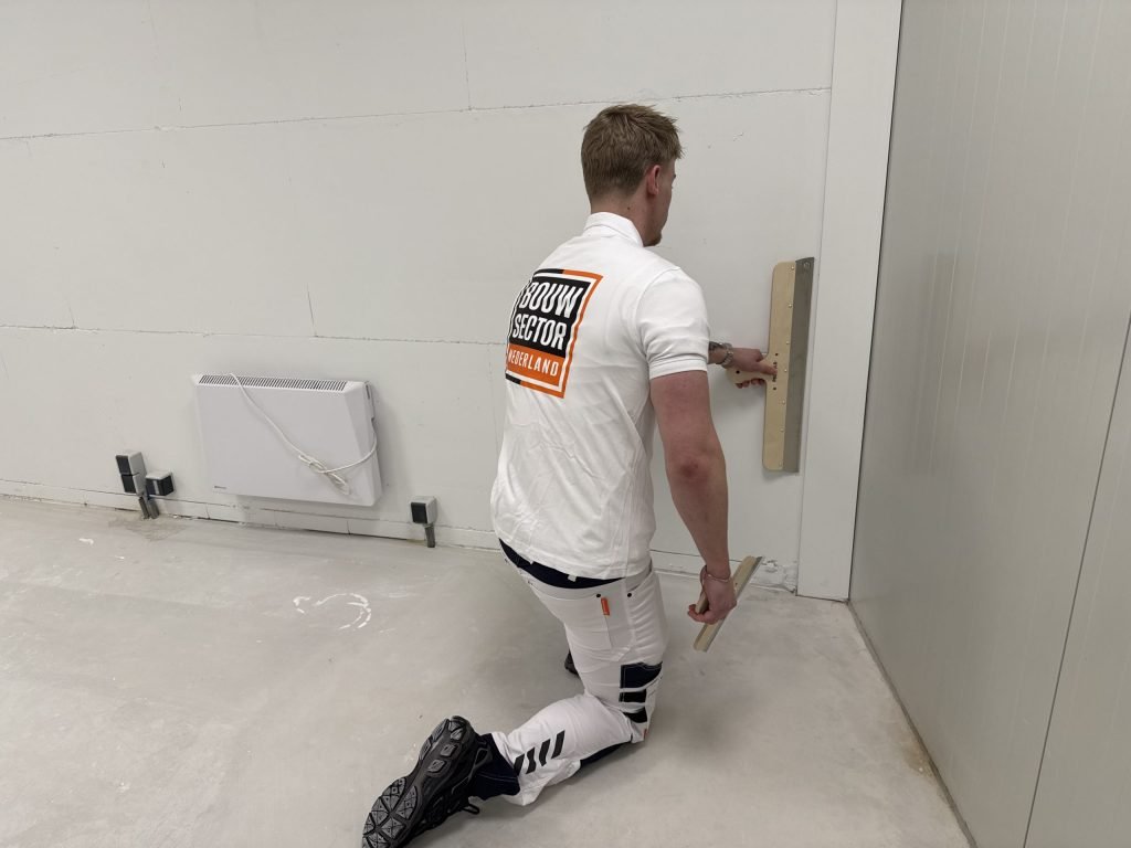 Stukadoor op een knie trekt een lange spaan langs de onderkant van een wand bij de vloer