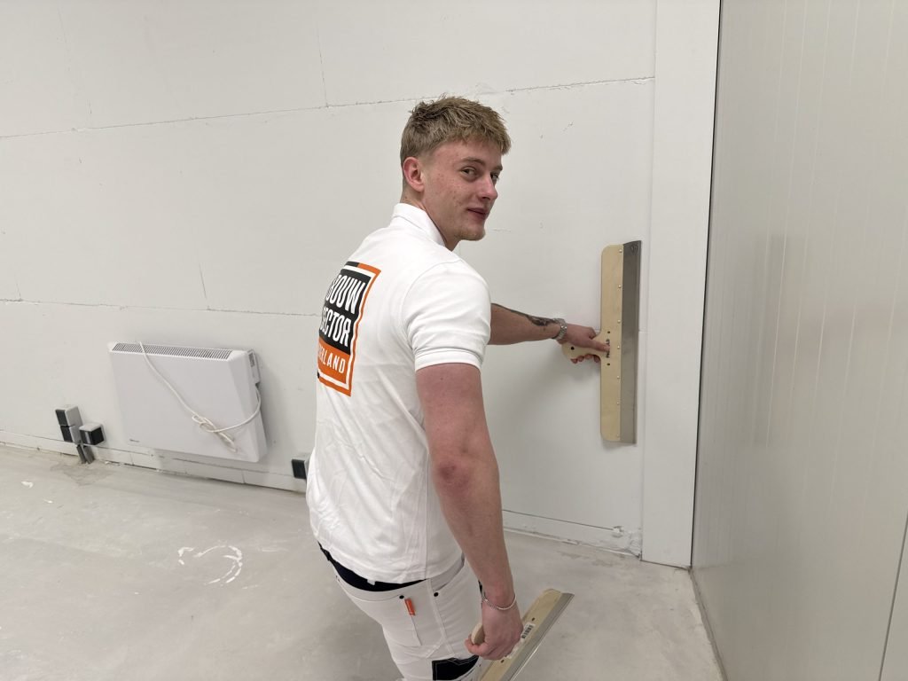 Stukadoor kijkt om terwijl hij met een lange spaan de onderkant van een witte wand afwerkt
