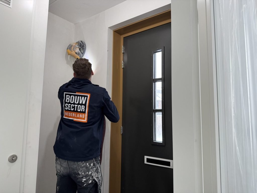 Vakman van Bouwsector Nederland in donkere werkjas schuurt de bovenkant van een wand bij de voordeur